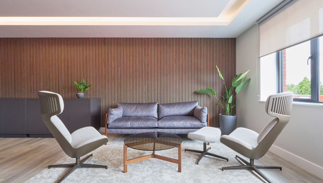 Modern lounge with a sleek gray sofa, two gray armchairs, and a round coffee table on a light rug. Wooden slat accent wall and large windows. Calm and inviting.