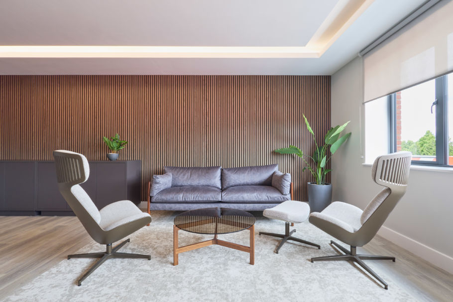Modern lounge with a sleek gray sofa, two gray armchairs, and a round coffee table on a light rug. Wooden slat accent wall and large windows. Calm and inviting.
