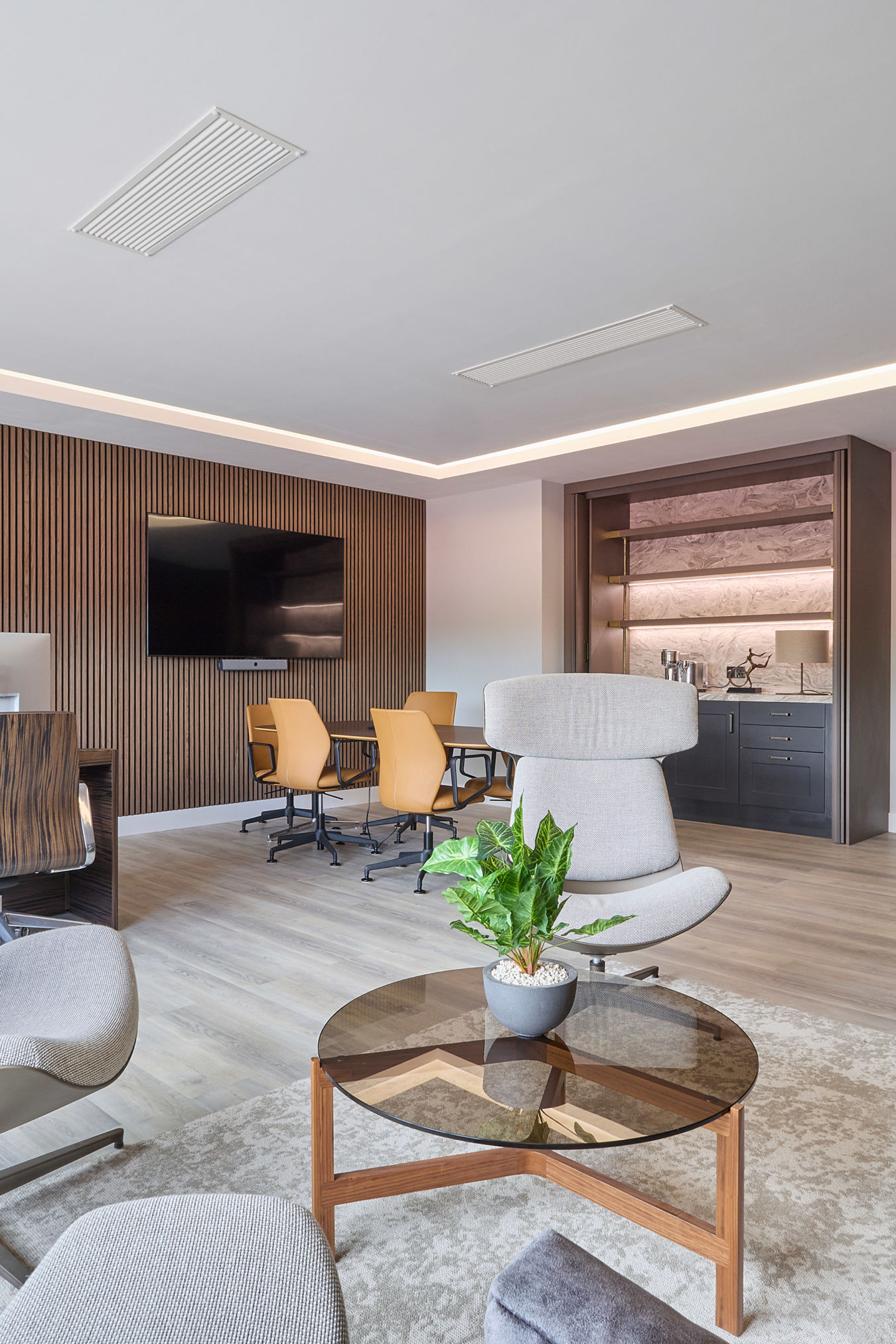 Modern office with a cozy ambiance, featuring plush gray chairs and a sofa around a glass table with a plant. Warm light, wood accents, and a TV on the wall add elegance.