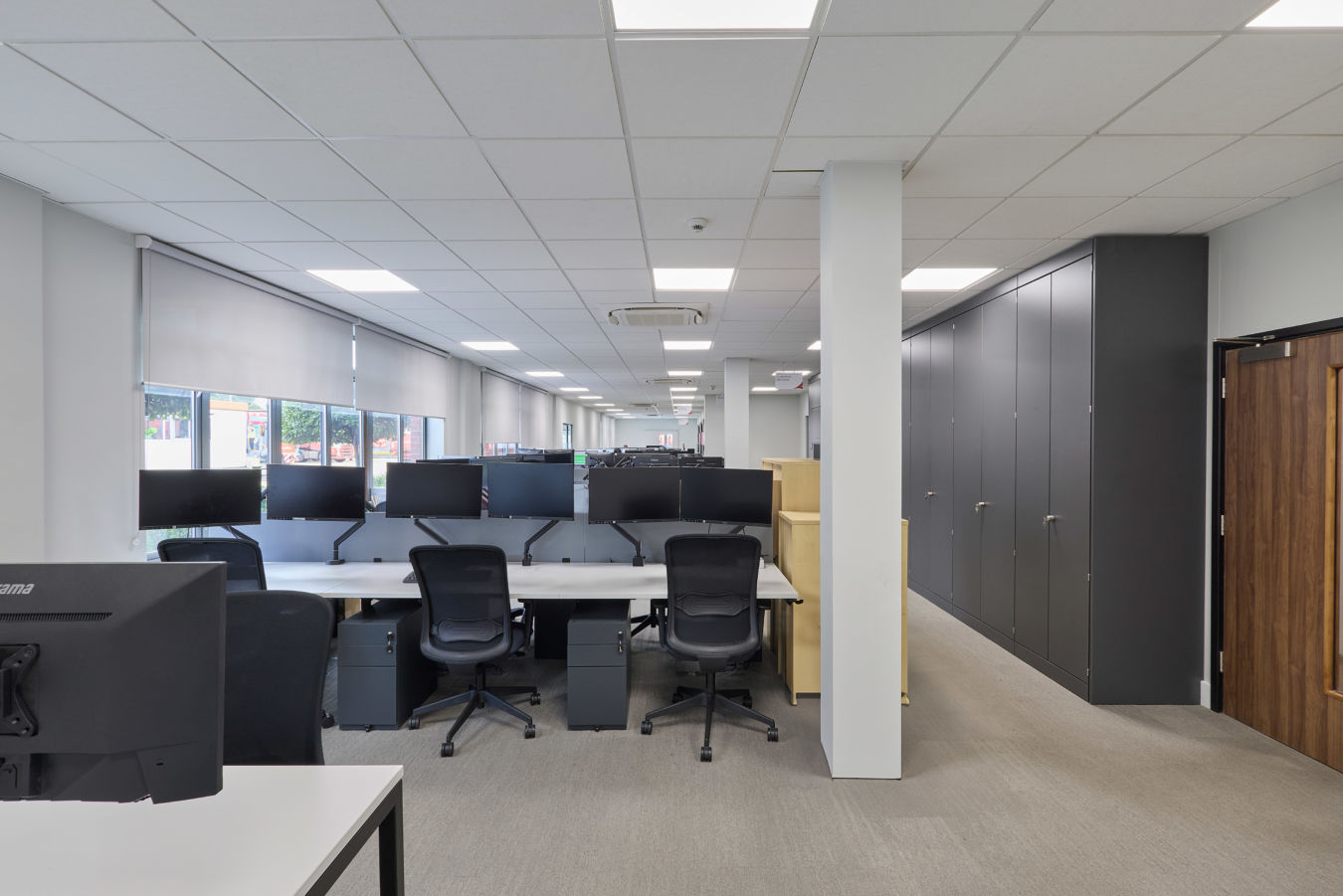 Wide-angle view of a modern, open-plan office with several desks, black chairs, aWide-angle view of a modern, open-plan office with several desks, black chairs, and multiple monitors. Neutral tones, well-lit, and organized.nd multiple monitors. Neutral tones, well-lit, and organized.