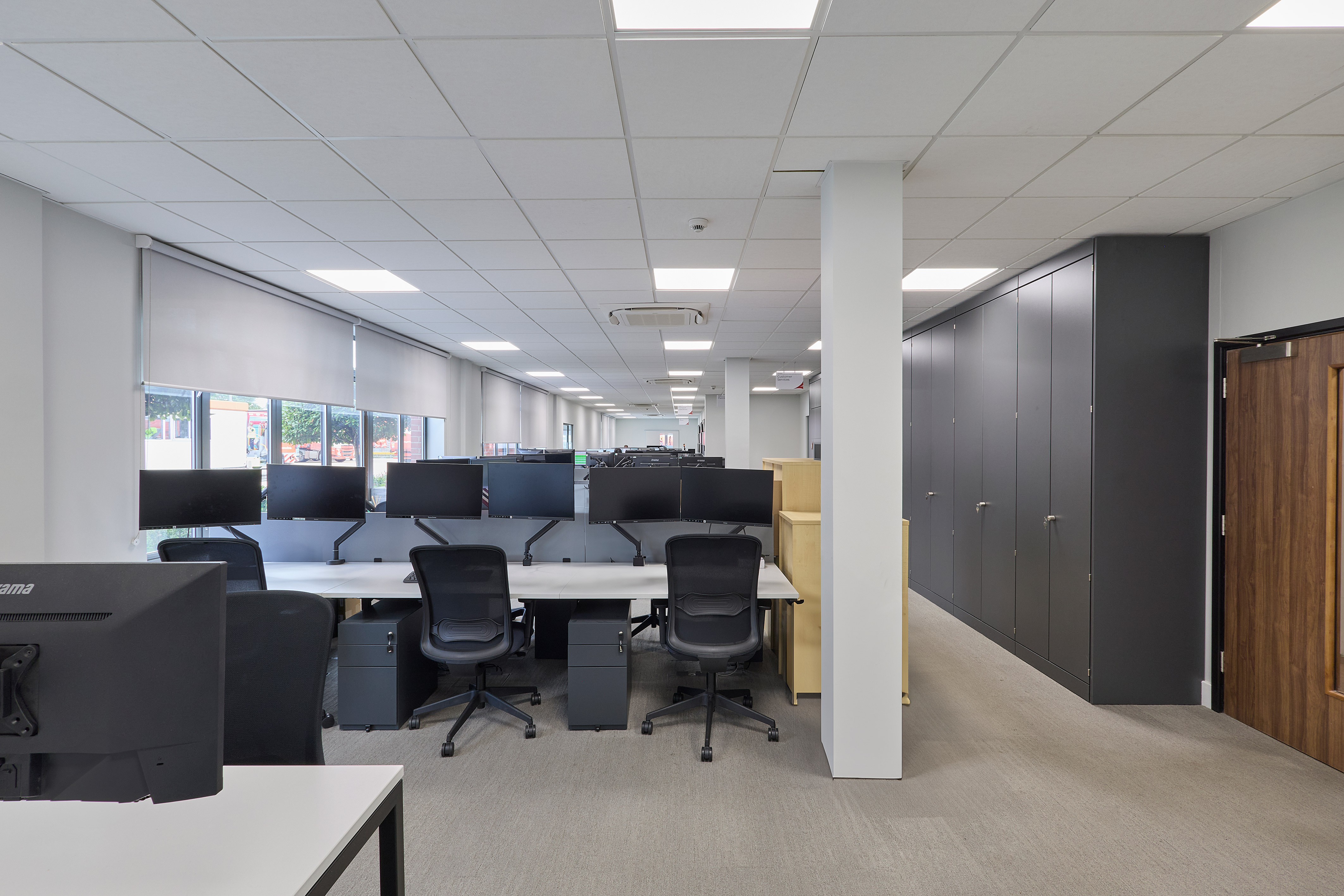 Wide-angle view of a modern, open-plan office with several desks, black chairs, aWide-angle view of a modern, open-plan office with several desks, black chairs, and multiple monitors. Neutral tones, well-lit, and organized.nd multiple monitors. Neutral tones, well-lit, and organized.