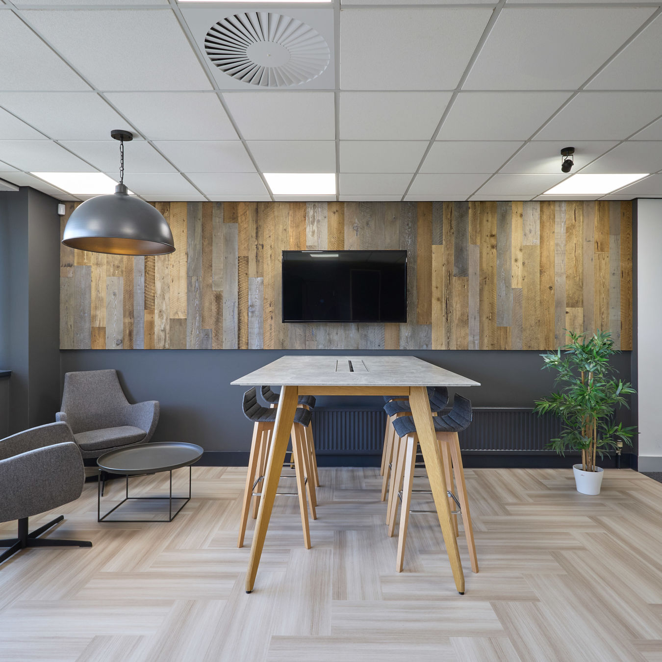Modern office lounge with wooden accent wall and mounted TV. Features a high table with stools, cozy armchairs, a small table, and a potted plant.
