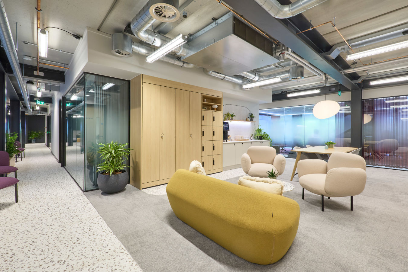 Modern office lounge with yellow sofa, beige chairs, and plants on gray carpet. Glass walls and exposed ducts create an open, airy feel.