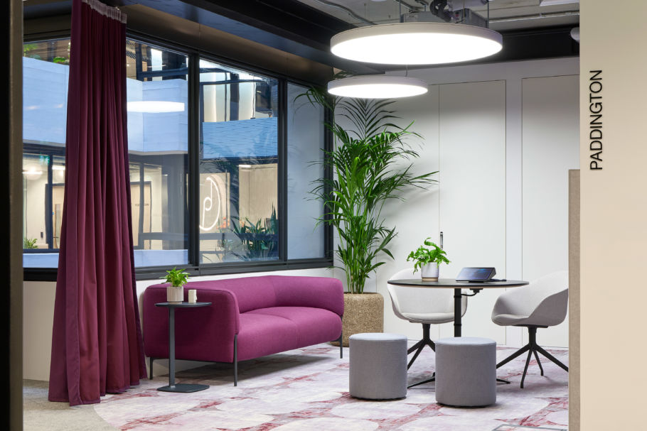 Modern office lounge with a magenta sofa, round table, and gray chairs. Large window and greenery add a fresh feel, softened by warm lighting.