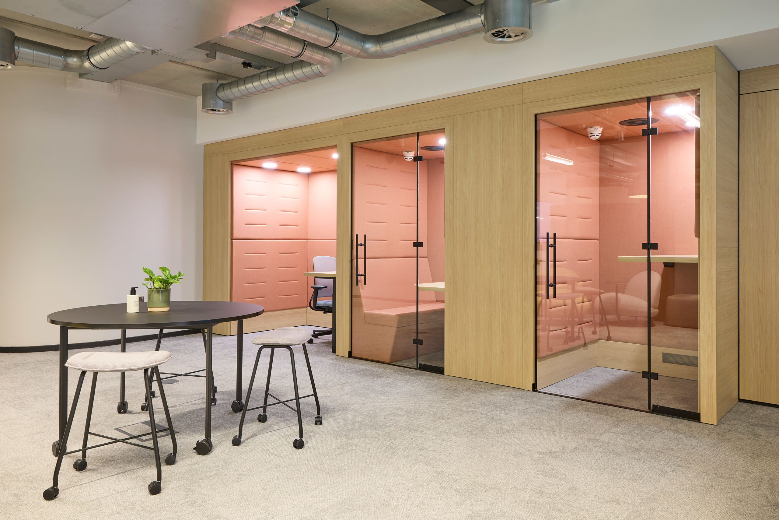 Modern office space with light wood and glass enclosed meeting pods, pink interiors. In front, a round table with stools and a plant. Industrial ceiling.