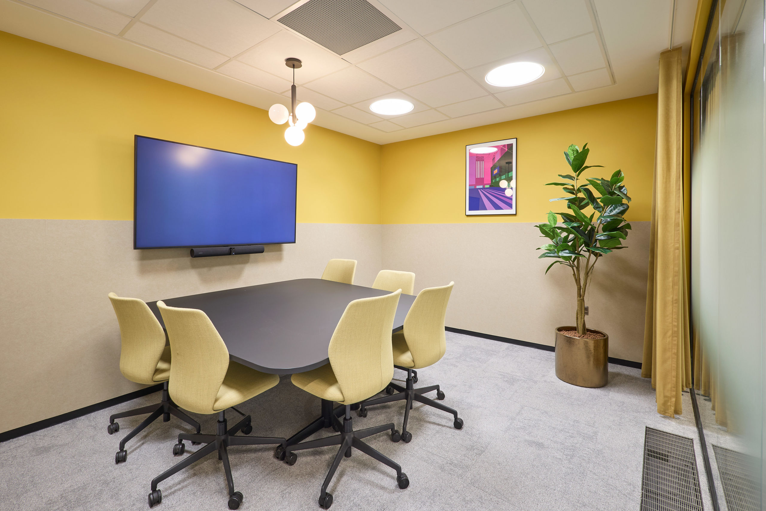 Modern meeting room with yellow walls, a large screen, and a black oval table surrounded by six beige chairs. A plant and abstract art add a welcoming touch.
