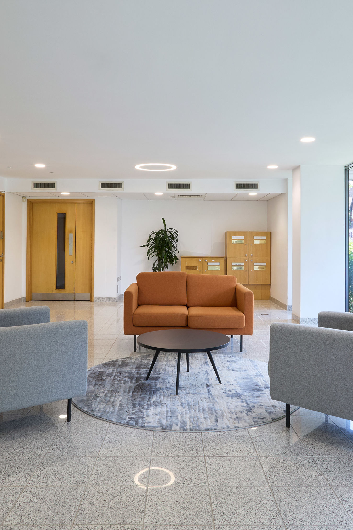 Modern office lobby with gray and orange sofas around a circular rug and table. Bright windows, plant, and wooden doors add a welcoming feel.