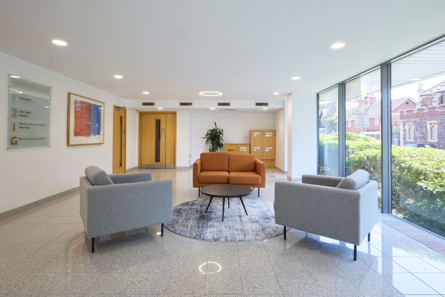 Modern office lobby with gray and orange sofas around a circular rug and table. Bright windows, plant, and wooden doors add a welcoming feel.