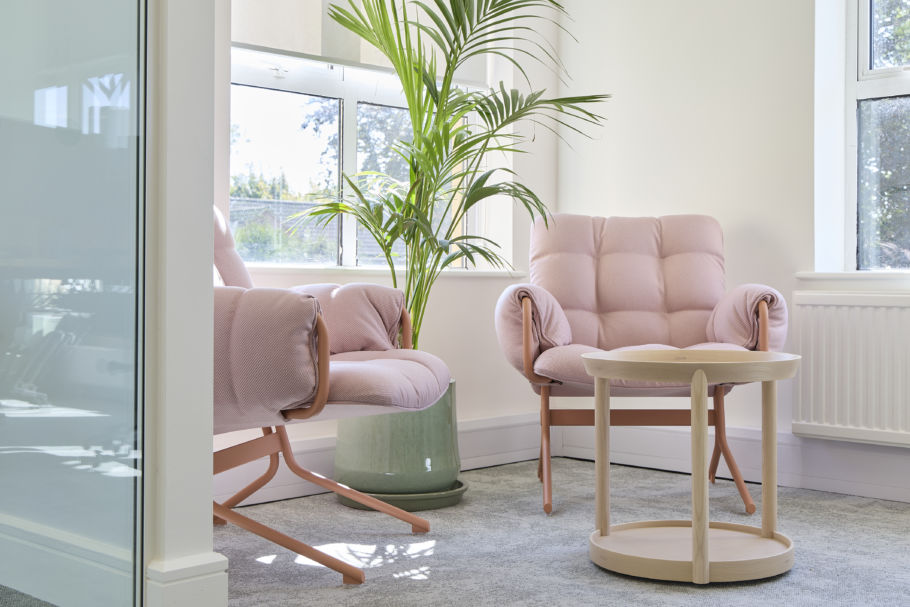 Bright, cozy room with two pink tufted armchairs, a round wooden table, and a tall green plant. Sunlight streams through large windows, creating a serene atmosphere.