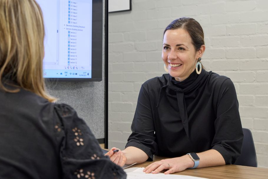 A female designer sitting at a boardroom table talking to a colleague with a screen to her right to show a briefing presentation